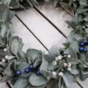 May include: A green and white wreath with blue berries. The wreath is made of faux greenery and is perfect for a holiday or winter decor.