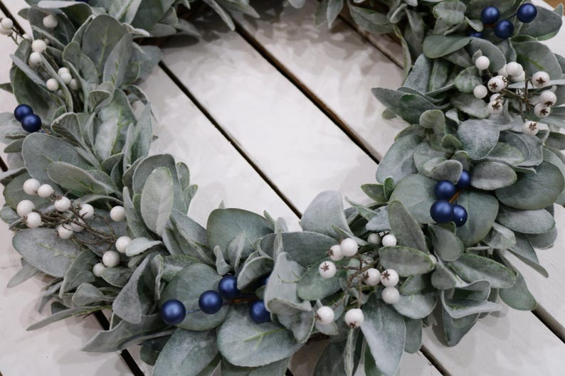 May include: A green and white wreath with blue berries. The wreath is made of faux greenery and is perfect for a holiday or winter decor.