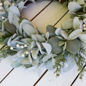 May include: A green and white artificial wreath with white pearl-like berries. The wreath is made of eucalyptus and cedar.