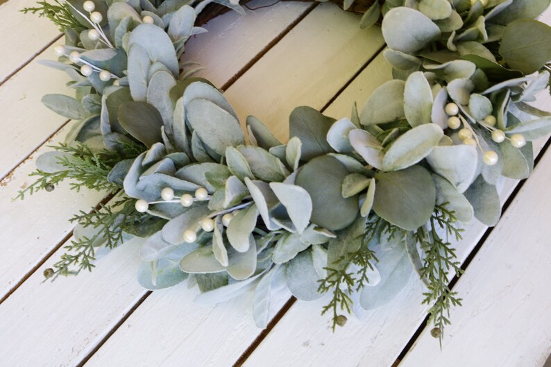 May include: A green and white artificial wreath with white pearl-like berries. The wreath is made of eucalyptus and cedar.