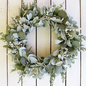 May include: A green and white eucalyptus wreath with white berries. The wreath is hanging on a white wooden background.