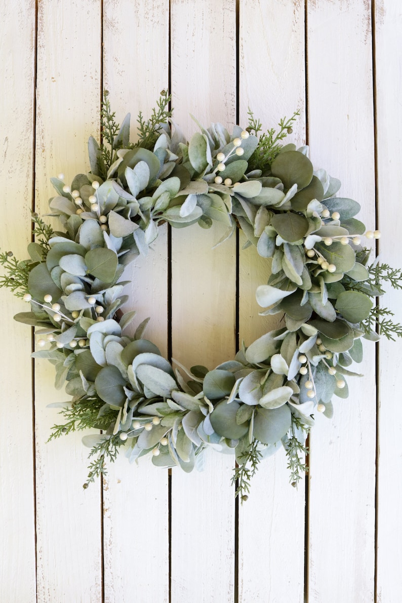 May include: A green and white eucalyptus wreath with white berries. The wreath is hanging on a white wooden background.