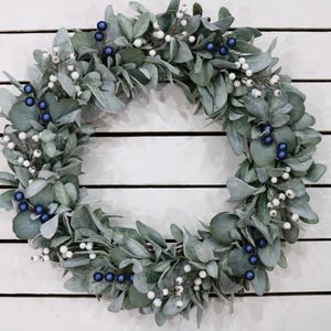 May include: A green wreath with white and blue berries. The wreath is made of artificial greenery and is perfect for decorating for the holidays.