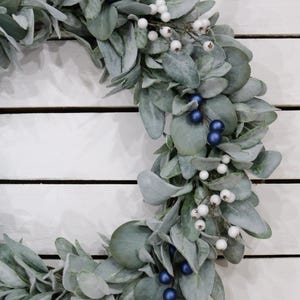 May include: A silver-green wreath with white berries and dark blue berries. The wreath is made of artificial greenery and is perfect for a winter or holiday decoration.