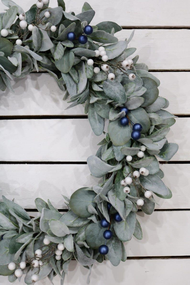 May include: A silver-green wreath with white berries and dark blue berries. The wreath is made of artificial greenery and is perfect for a winter or holiday decoration.
