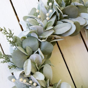 May include: A green and white faux eucalyptus wreath with white pearl-like berries. The wreath is made of artificial greenery and is perfect for a farmhouse or rustic decor style.