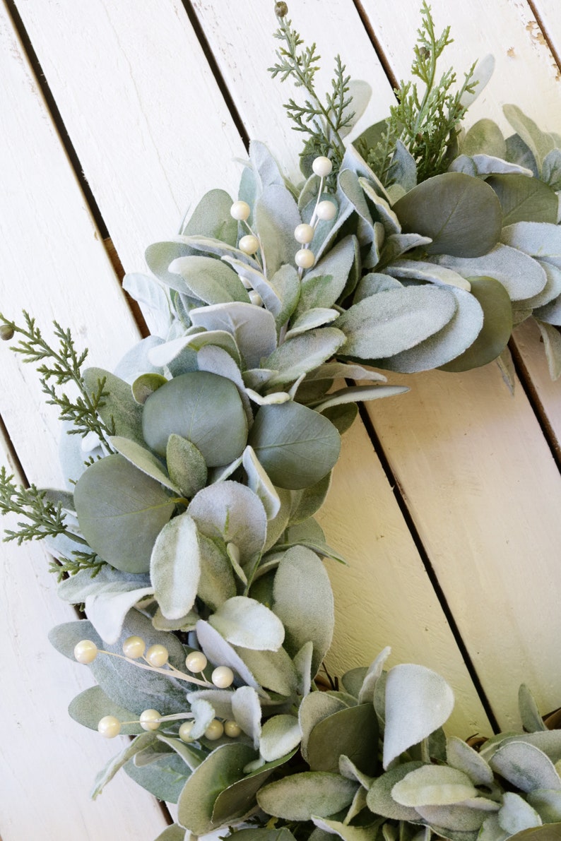 May include: A green and white faux eucalyptus wreath with white pearl-like berries. The wreath is made of artificial greenery and is perfect for a farmhouse or rustic decor style.