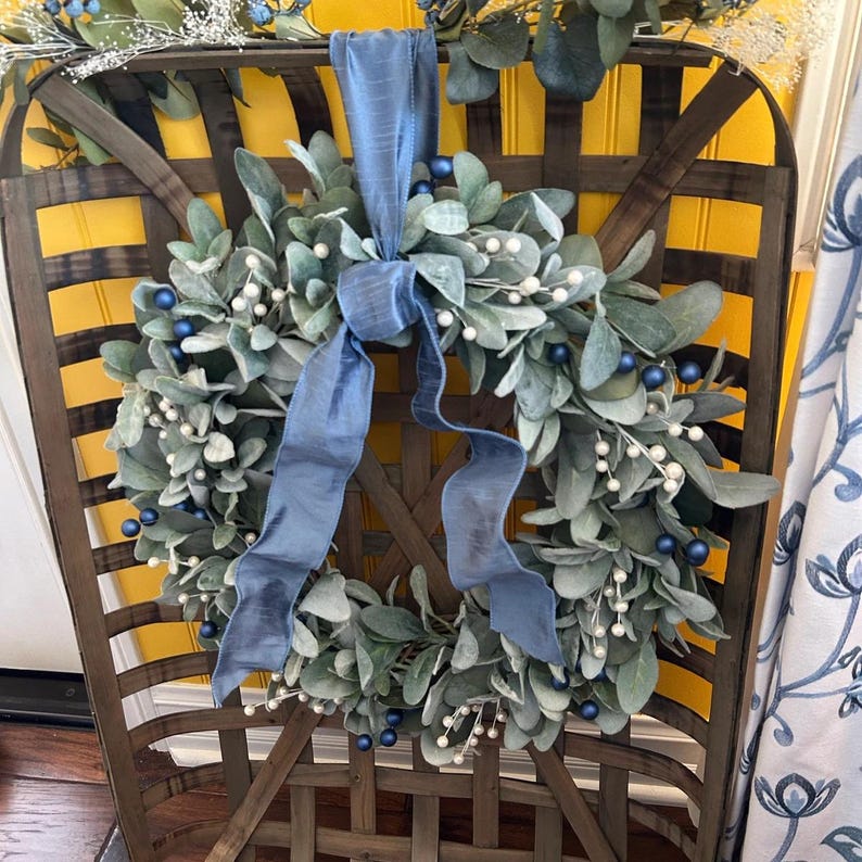 May include: A blue and white wreath with a blue ribbon hanging on a wooden basket. The wreath is made of eucalyptus leaves and has white berries and blue ornaments.
