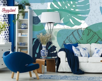 Può includere: Un divano bianco con cuscini e una coperta blu si trova in un soggiorno con una carta da parati a motivi di foglie tropicali blu e verdi. Una poltrona blu con gambe in legno si trova in primo piano. Una lampada bianca con un piedistallo nero si trova sullo sfondo.