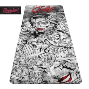 May include: A black and white yoga mat with a graphic design featuring a skull, a woman with a sugar skull face, and dollar bills. The text "Gaby" is written in red at the top of the mat.