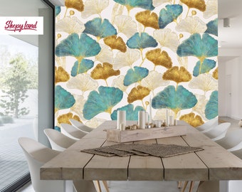 Op de afbeelding: Behang met een herhalend patroon van gestileerde ginkgo bladeren in teal, goud en gebroken wit. De bladeren zijn geplaatst tegen een lichte achtergrond. De kamer heeft een houten eettafel en witte stoelen.