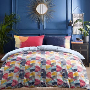 Op de afbeelding: Een kingsize bed met een kleurrijk geometrisch patroon dekbedovertrek. Het dekbedovertrek heeft een patroon van overlappende cirkels in tinten roze, geel, blauw en grijs. Het bed is opgemaakt met een lichtblauw laken en twee kussens, een rood en een donkerblauw met witte stippen. De tekst "KING SIZE PREVIEW" is zichtbaar onderaan de afbeelding.