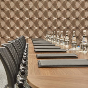 Op de afbeelding: Een lange houten vergadertafel ingericht met meerdere zwarte lederen mappen en doorzichtige glazen waterflessen. Een rij zwarte bureaustoelen met gaasrugleuning staat opgesteld langs de tafel, die voor een muur met geometrisch patroon staat.