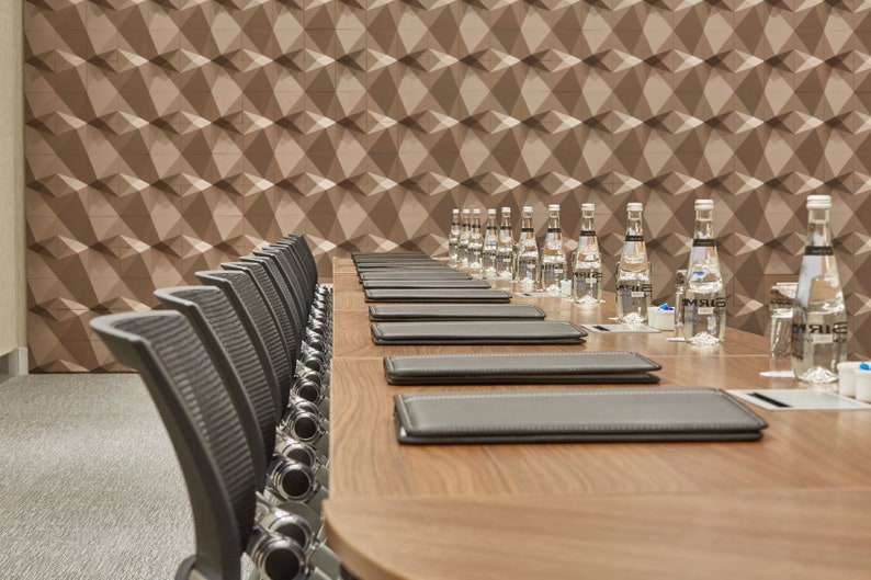 Op de afbeelding: Een lange houten vergadertafel ingericht met meerdere zwarte lederen mappen en doorzichtige glazen waterflessen. Een rij zwarte bureaustoelen met gaasrugleuning staat opgesteld langs de tafel, die voor een muur met geometrisch patroon staat.