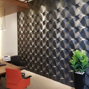 Op de afbeelding: Een muur bedekt met donkergrijze, driedimensionale geometrische panelen met een gefacetteerd, driehoekig patroon. De muur heeft een herhalend hoekig ontwerp dat diepte en schaduw cre&euml;ert, verlicht door plafondverlichting in een kantoor.