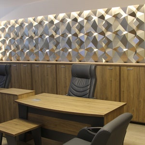 Op de afbeelding: Een kantoorinterieur met twee houten bureaus, donkergrijze stoelen en bijpassende houten opbergkasten. Boven de kasten bevindt zich een wand met een geometrisch, driedimensionaal paneelontwerp dat wordt verlicht door ge&iuml;ntegreerde verlichting.