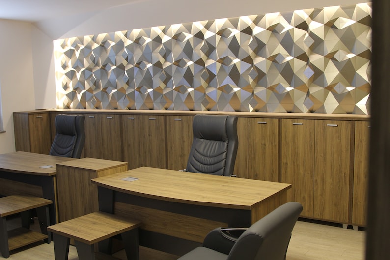 Op de afbeelding: Een kantoorinterieur met twee houten bureaus, donkergrijze stoelen en bijpassende houten opbergkasten. Boven de kasten bevindt zich een wand met een geometrisch, driedimensionaal paneelontwerp dat wordt verlicht door ge&iuml;ntegreerde verlichting.