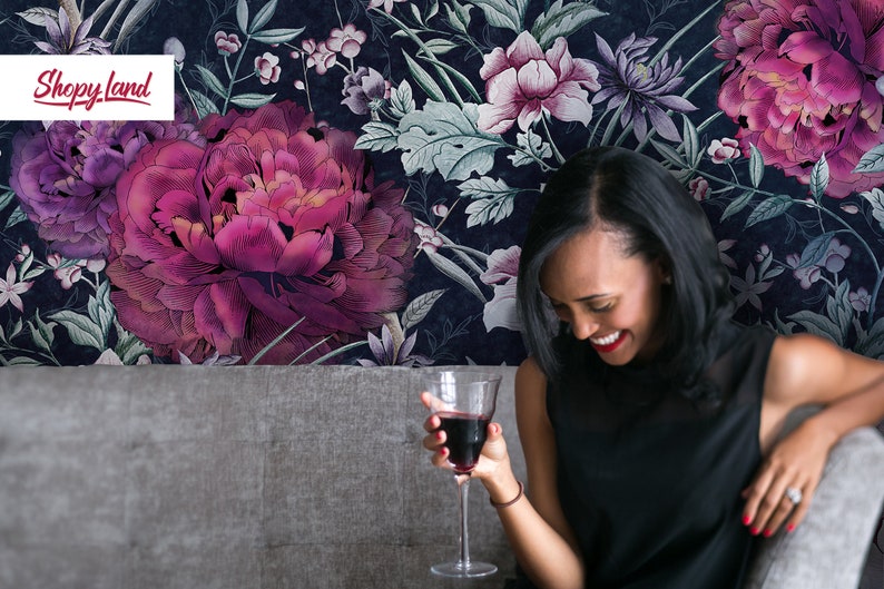 May include: A woman smiles while holding a glass of red wine, sitting on a gray couch in front of a dark blue floral wallpaper with large pink flowers.