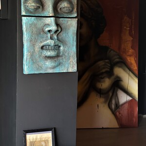 May include: A teal-colored, two-piece wall sculpture of a human face with closed eyes and open mouth. A framed portrait and a large painting of a figure are in the background. A cat rests on a fluffy rug.