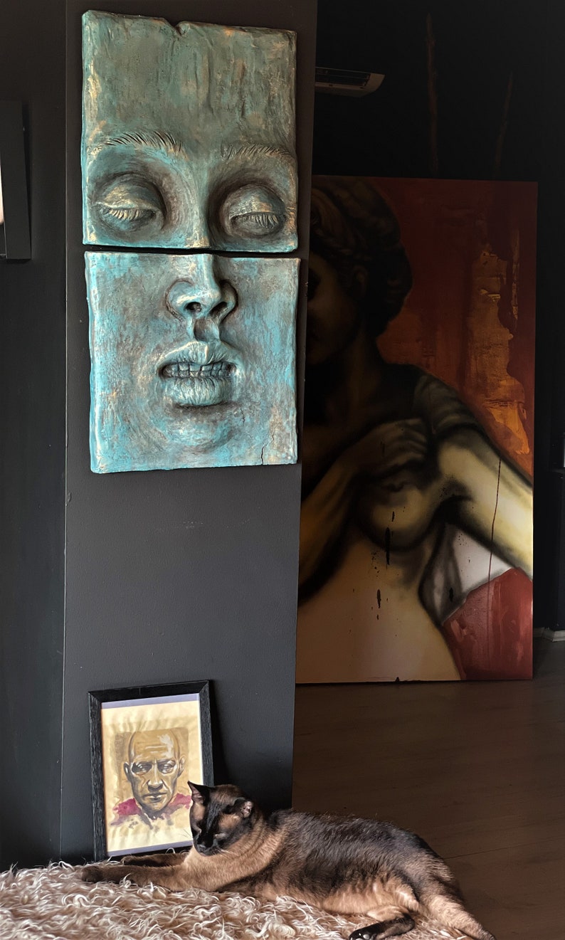 May include: A teal-colored, two-piece wall sculpture of a human face with closed eyes and open mouth. A framed portrait and a large painting of a figure are in the background. A cat rests on a fluffy rug.