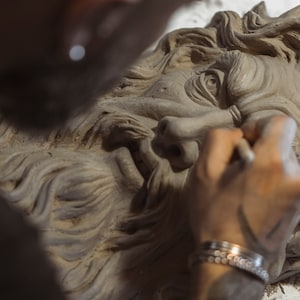 May include: A person sculpting a detailed clay bust of a bearded man with a serious expression. The bust is unfinished, with the artist's hand visible as they work on the face.