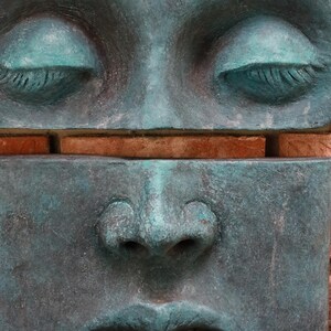 May include: A close-up of a weathered, teal-colored sculpture of a face with closed eyes and a serene expression. The sculpture is divided horizontally, with a section of brick visible between the upper and lower halves.