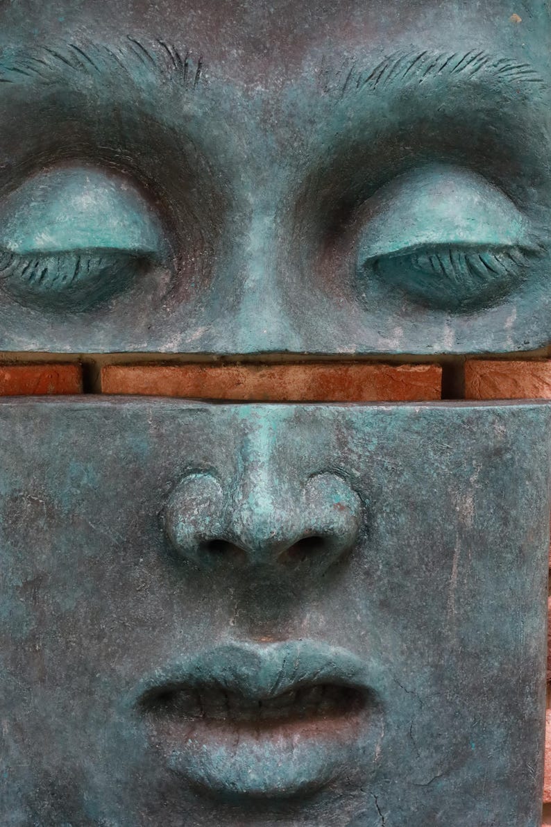 May include: A close-up of a weathered, teal-colored sculpture of a face with closed eyes and a serene expression. The sculpture is divided horizontally, with a section of brick visible between the upper and lower halves.