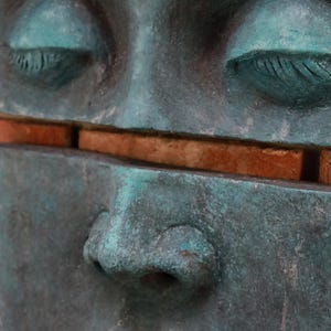 May include: Close-up of a weathered teal-colored sculpture of a human face with closed eyes. A horizontal gap reveals a section of reddish-brown brick. The sculpture's surface has a textured, aged appearance.