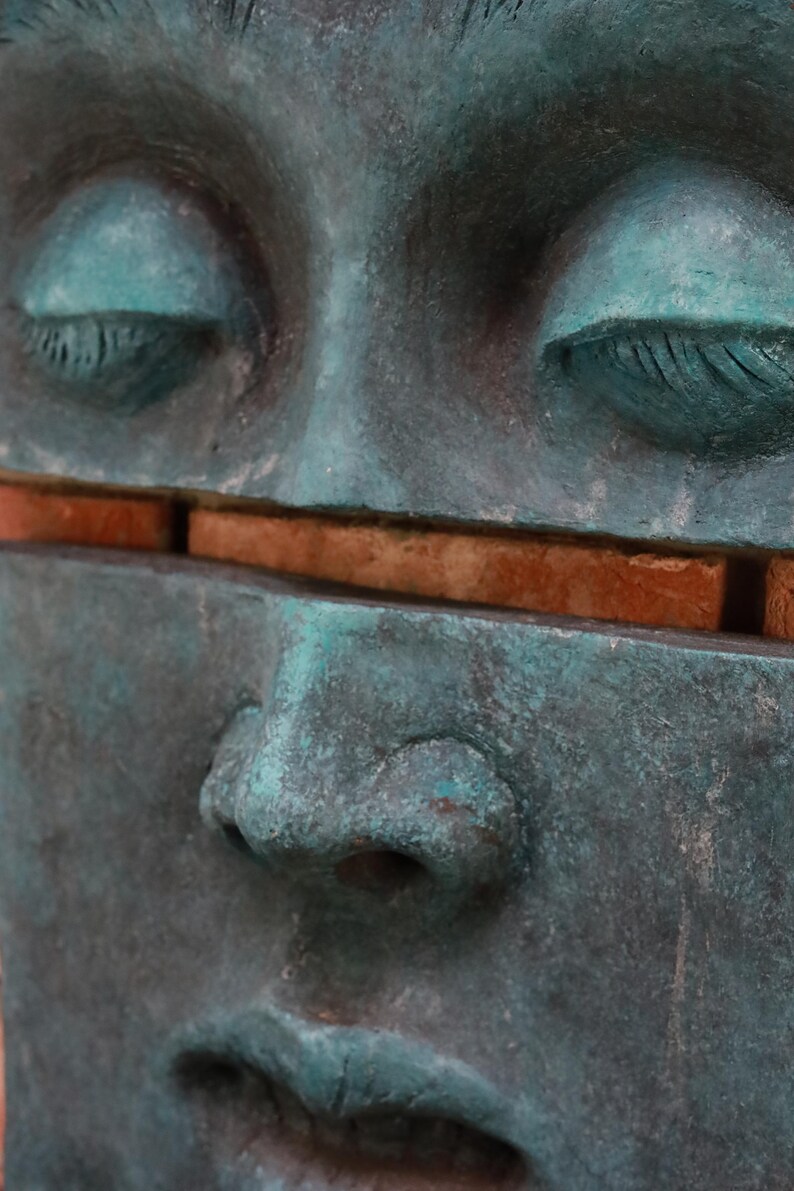 May include: Close-up of a weathered teal-colored sculpture of a human face with closed eyes. A horizontal gap reveals a section of reddish-brown brick. The sculpture's surface has a textured, aged appearance.