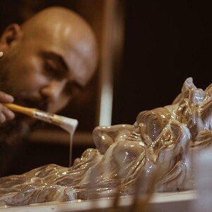 May include: A person is painting a detailed, white, ceramic sculpture of a mythical creature with a brush. The sculpture has a long beard and horns.
