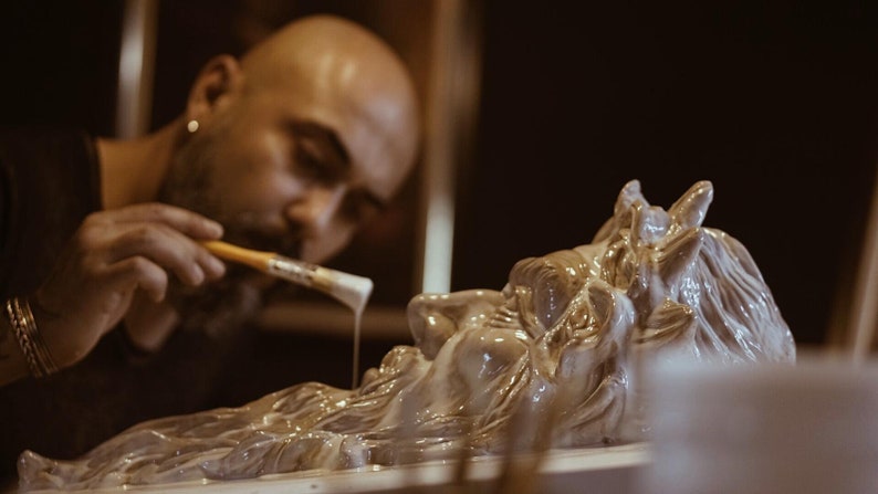 May include: A person is painting a detailed, white, ceramic sculpture of a mythical creature with a brush. The sculpture has a long beard and horns.