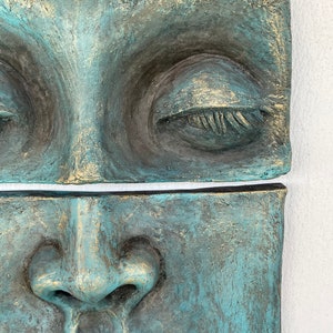 May include: Close-up of a two-part sculpture depicting a human face. The sculpture has a textured surface with a teal and gold color scheme. The top section shows closed eyes, while the bottom section shows the nose and mouth.