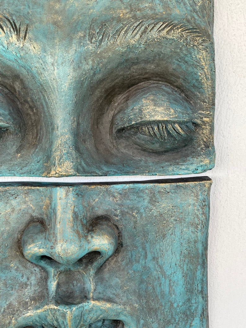 May include: Close-up of a two-part sculpture depicting a human face. The sculpture has a textured surface with a teal and gold color scheme. The top section shows closed eyes, while the bottom section shows the nose and mouth.