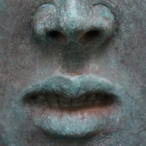 May include: Close-up of a weathered, square-shaped sculpture of a face. The nose and lips are prominent, with a turquoise-green patina contrasting against the darker, textured surface. The sculpture has a slightly aged appearance.