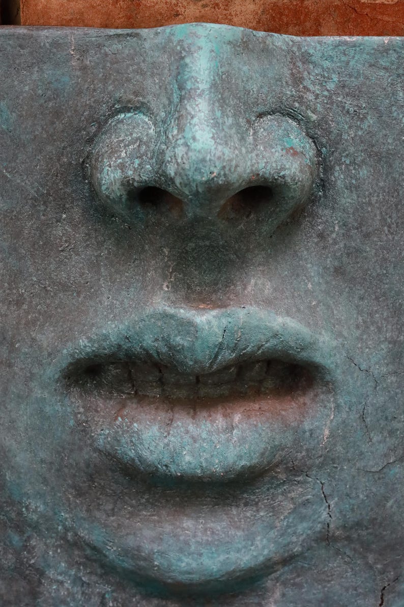 May include: Close-up of a weathered, square-shaped sculpture of a face. The nose and lips are prominent, with a turquoise-green patina contrasting against the darker, textured surface. The sculpture has a slightly aged appearance.