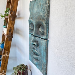 May include: A two-piece wall sculpture depicting a human face with closed eyes and parted lips. The artwork is a weathered teal color with gold accents, mounted on a white wall. The sculpture has a textured surface and a rustic aesthetic.