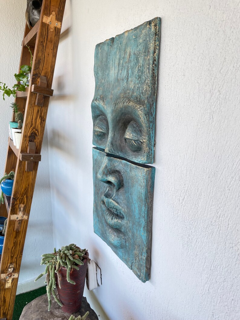 May include: A two-piece wall sculpture depicting a human face with closed eyes and parted lips. The artwork is a weathered teal color with gold accents, mounted on a white wall. The sculpture has a textured surface and a rustic aesthetic.