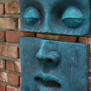 May include: A teal-colored, sculpted face composed of two rectangular sections, mounted on a brick wall. The upper section features closed eyes with detailed eyelashes, while the lower section displays a nose and lips. The sculpture has a weathered appearance.