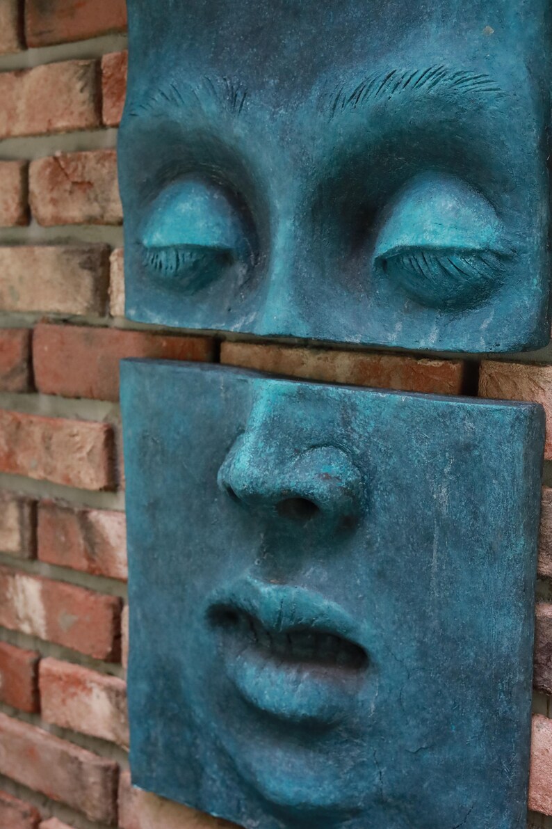 May include: A teal-colored, sculpted face composed of two rectangular sections, mounted on a brick wall. The upper section features closed eyes with detailed eyelashes, while the lower section displays a nose and lips. The sculpture has a weathered appearance.