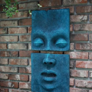 May include: Two square, teal-colored wall sculptures depicting a human face. The top sculpture shows closed eyes, and the bottom shows the nose and mouth. The sculptures are mounted on a brick wall.
