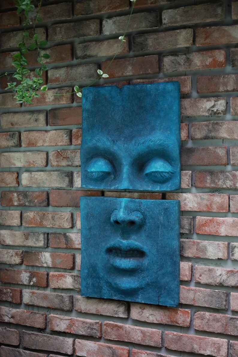May include: Two square, teal-colored wall sculptures depicting a human face. The top sculpture shows closed eyes, and the bottom shows the nose and mouth. The sculptures are mounted on a brick wall.