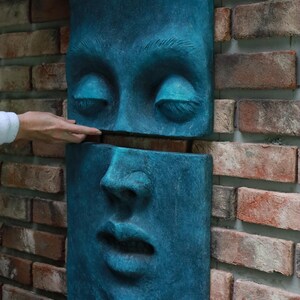 May include: A teal-colored, two-piece wall sculpture depicting a human face. The top section shows closed eyes, while the bottom section displays the nose and mouth. The sculpture is mounted on a brick wall.