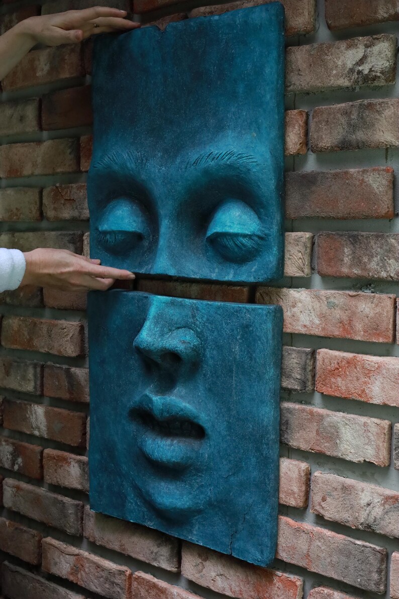 May include: A teal-colored, two-piece wall sculpture depicting a human face. The top section shows closed eyes, while the bottom section displays the nose and mouth. The sculpture is mounted on a brick wall.