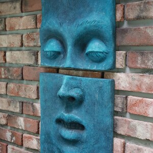 May include: Two-piece wall art featuring a face in a teal blue hue. The top piece shows closed eyes, and the bottom piece depicts the nose and mouth. The art is mounted on a brick wall, creating a striking contrast.