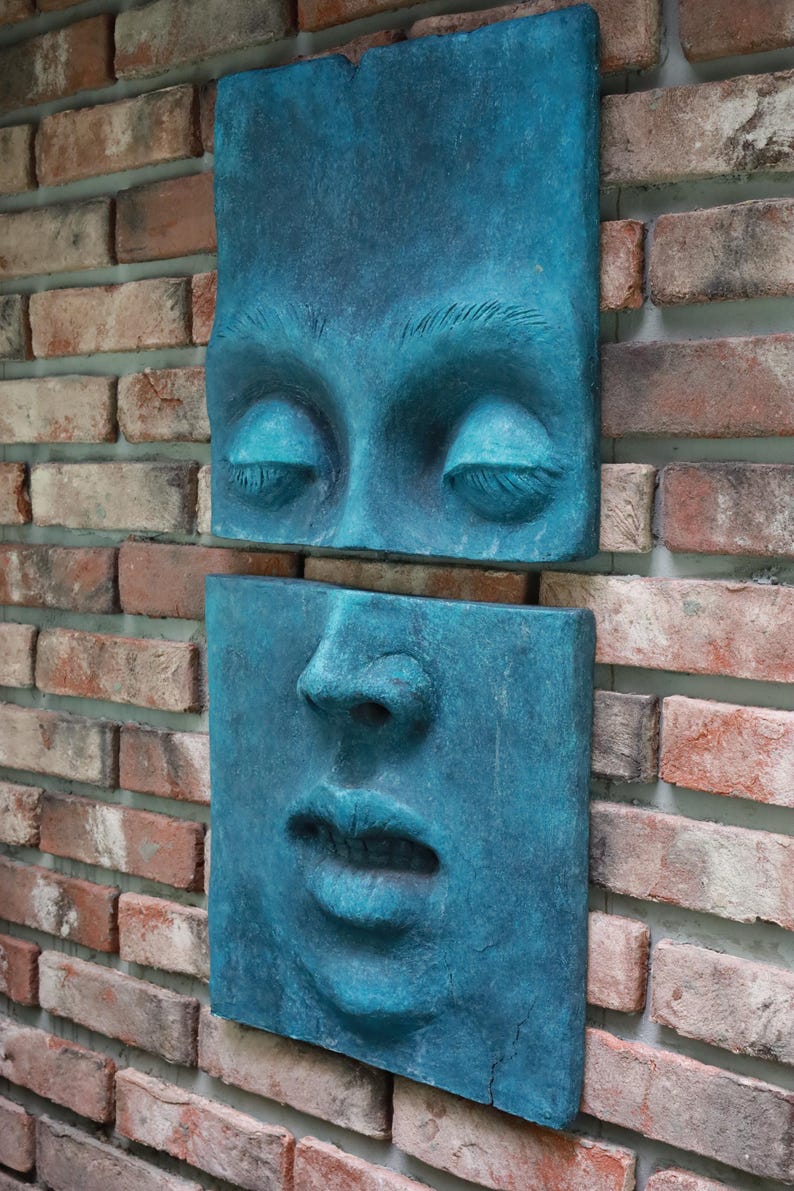 May include: Two-piece wall art featuring a face in a teal blue hue. The top piece shows closed eyes, and the bottom piece depicts the nose and mouth. The art is mounted on a brick wall, creating a striking contrast.