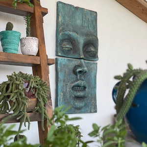 May include: A teal-colored wall sculpture depicting a human face, split into two rectangular sections. The eyes are closed, and the lips are slightly parted. The sculpture is mounted on a white wall, with a wooden ladder shelf holding potted plants in the background.