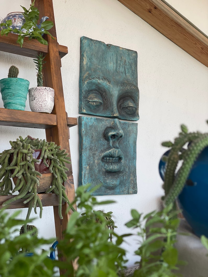 May include: A teal-colored wall sculpture depicting a human face, split into two rectangular sections. The eyes are closed, and the lips are slightly parted. The sculpture is mounted on a white wall, with a wooden ladder shelf holding potted plants in the background.