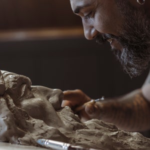 May include: A bearded artist carefully sculpts a clay bust of a person's face. The artist is using a small tool to shape the clay.