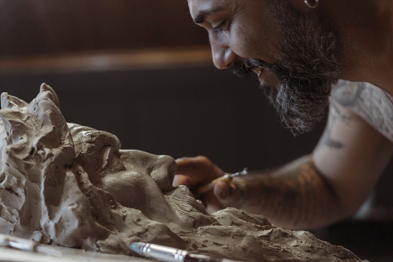 May include: A bearded artist carefully sculpts a clay bust of a person's face. The artist is using a small tool to shape the clay.