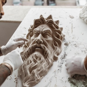 May include: A person is working on a clay sculpture of a man's face with a long beard. The sculpture is on a white surface with some clay debris. The person is wearing white gloves.